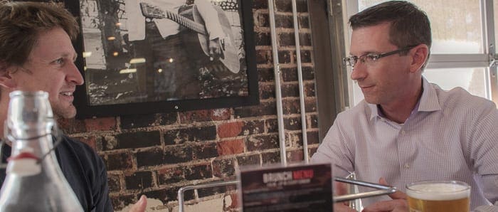 Two men sitting at a table indoors with drinks having a conversation