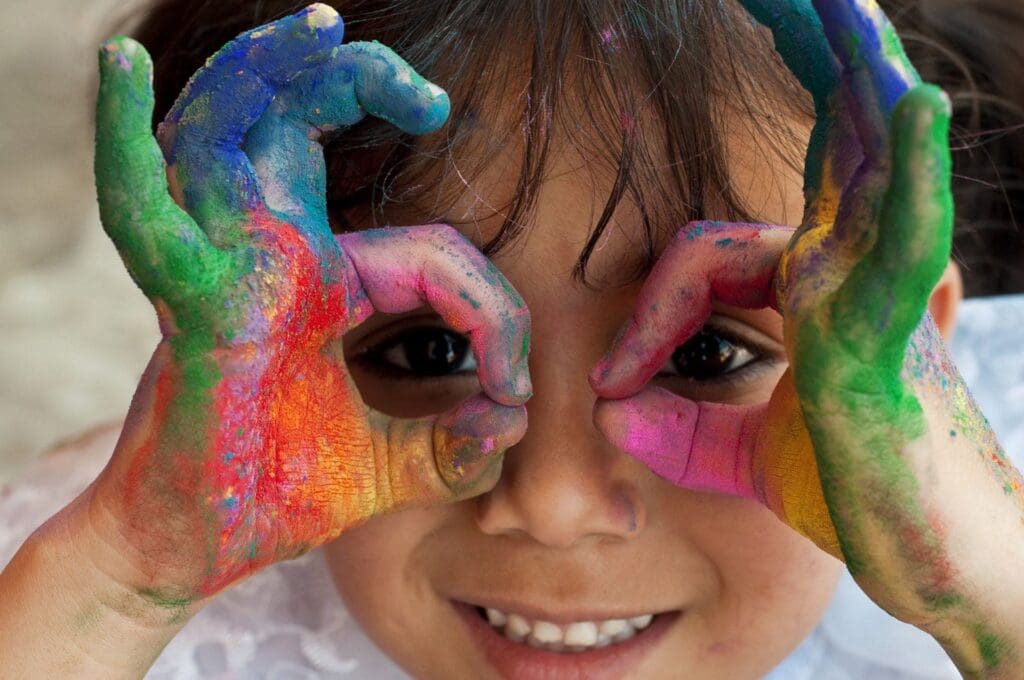young child with multicolored paint on their hands making circles around their eyes with their fingers